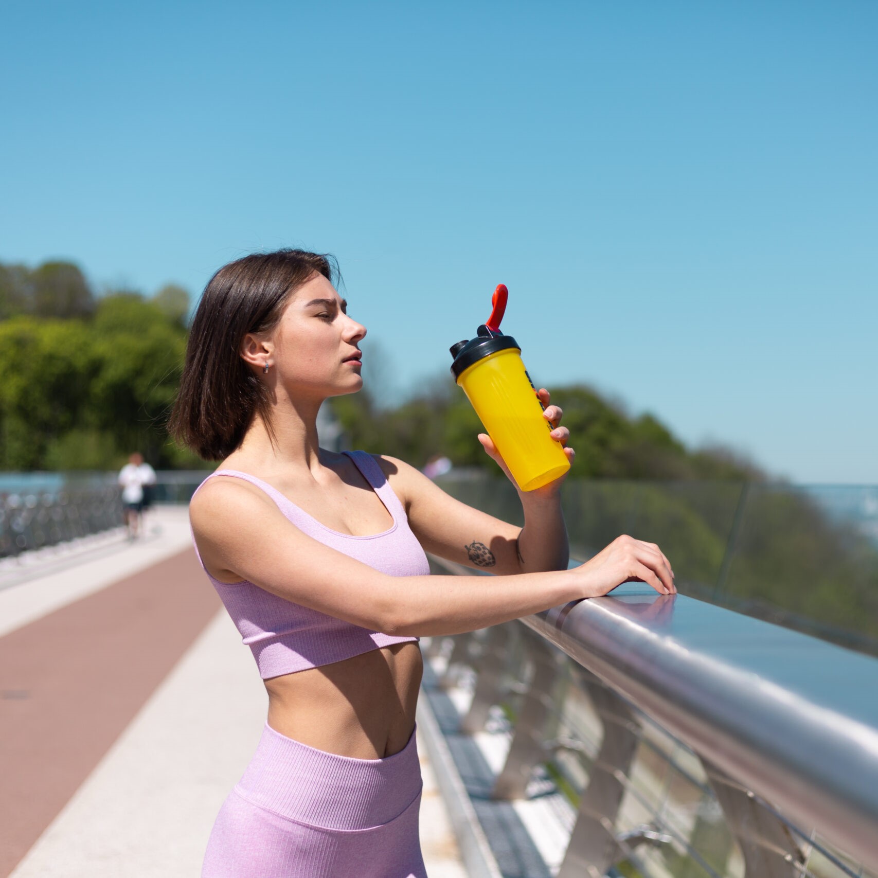 Young woman in fitting sport wear on bridge at hot sunny morning with bottle of water shaker thirsty after workout tired drinking